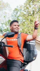 Entregador sorridente em motocicleta fazendo gesto com a mão, representando trabalho por aplicativo como renda extra